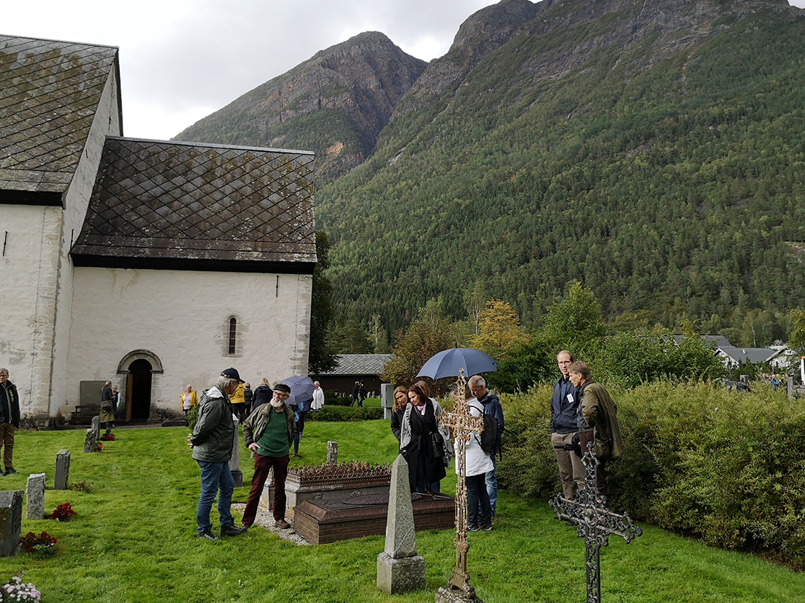 Folk står samlet rundt en gammel grav utenfor en hvit kirke med høye fjell i bakgrunnen.