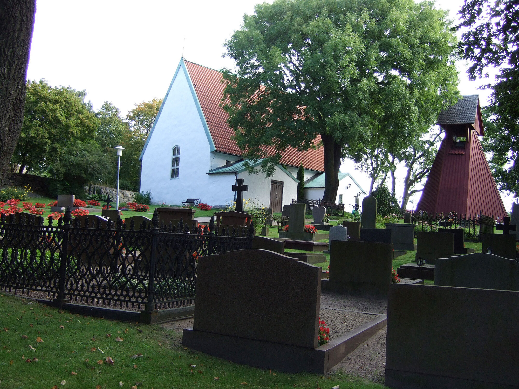 Hvit kirke av mur med rødt tak med en kirkegård og et stort løvtre utenfor.